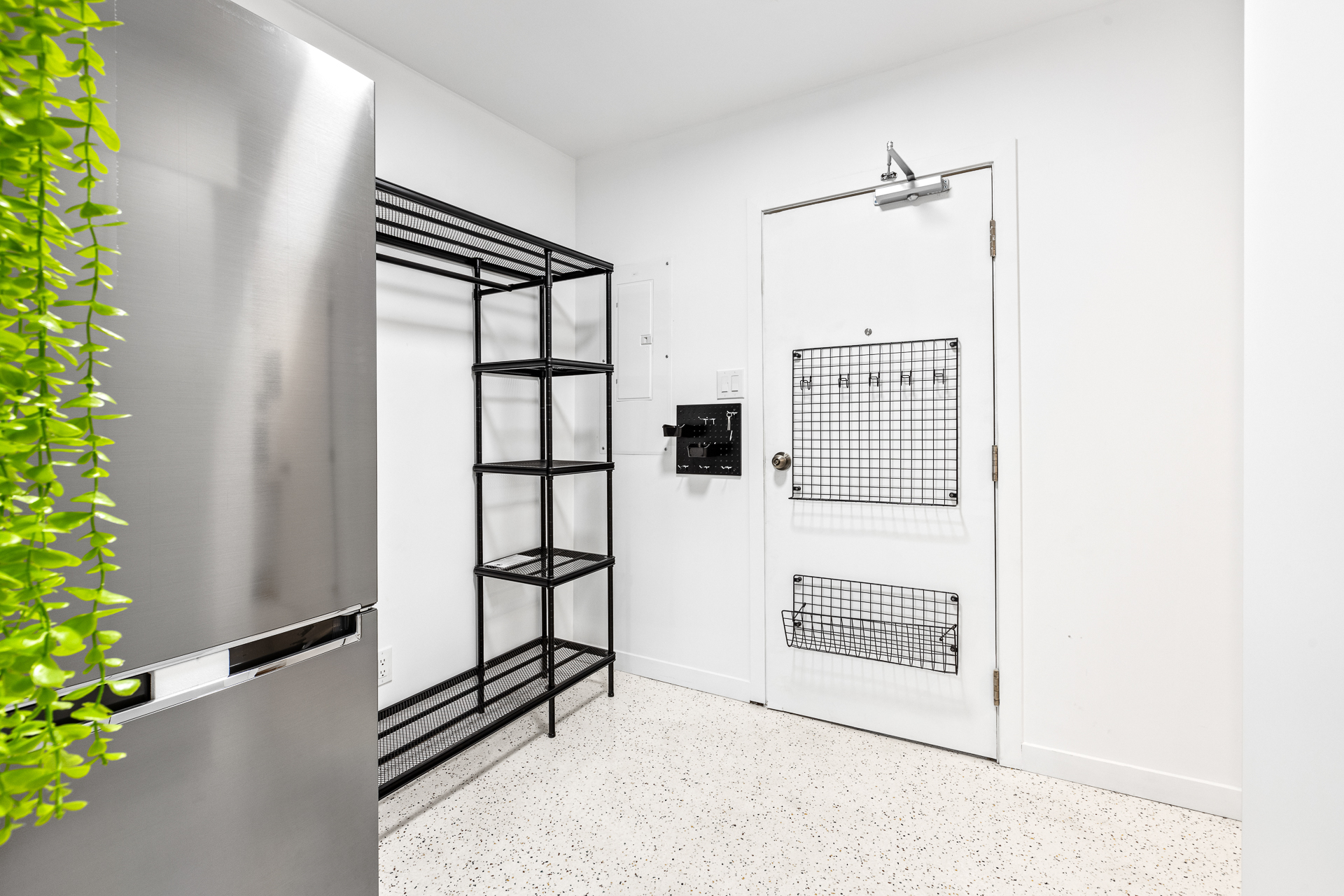 Unit 09 entry zone: heavy-duty steel wardrobe with top luggage shelf, side shelves, base shelf, and full-width steel rod; LG full-size refrigerator on the kitchen wall behind a slim divider; speckled terrazzo-style floor; in-suite laundry visible at the kitchen wall.