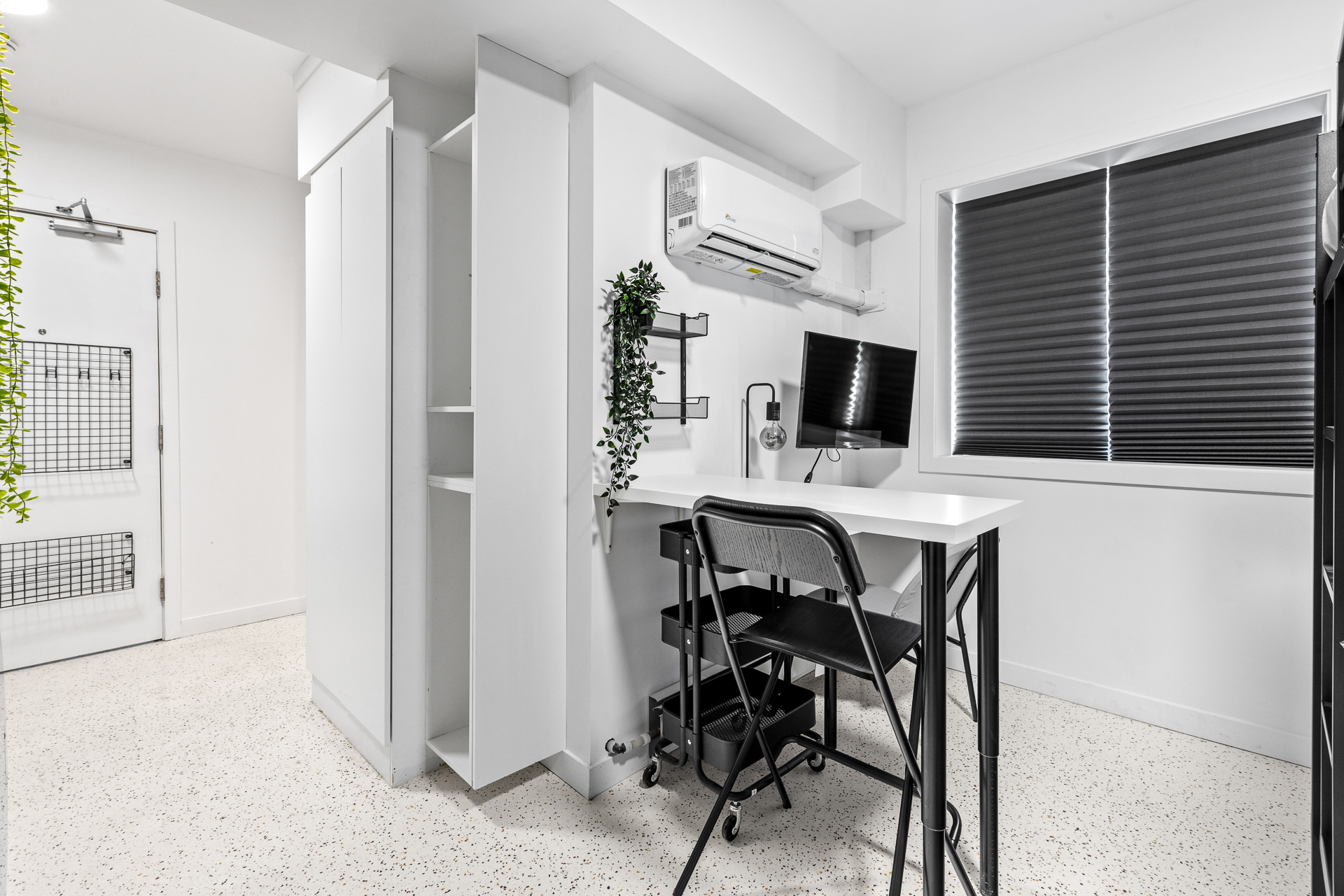 Unit 09 desk wall: wall-mounted desk with task chair under a flat-screen TV, perpendicular bar-top with stool, acacia-base task lamp, private mini-split HVAC, blackout shade, and a two-tier steel shelf.