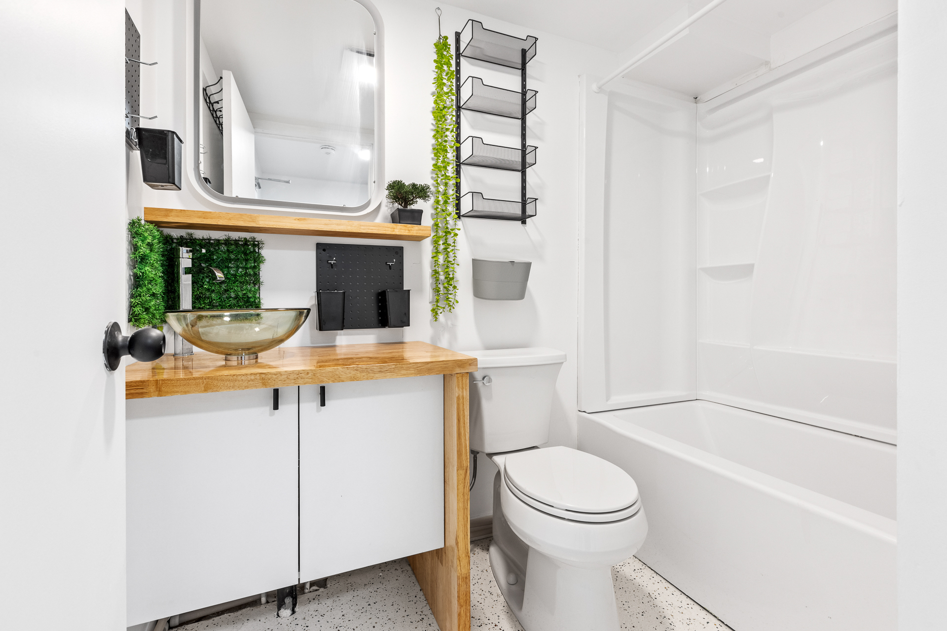 Unit 09 bathroom: butcher-block vanity with glass vessel sink and white cabinet base, full-height rounded mirror with floating shelf, magnetic accessory boards on the wall, wall-mounted wire organizer ladder, entry coat rack, and toilet at right.